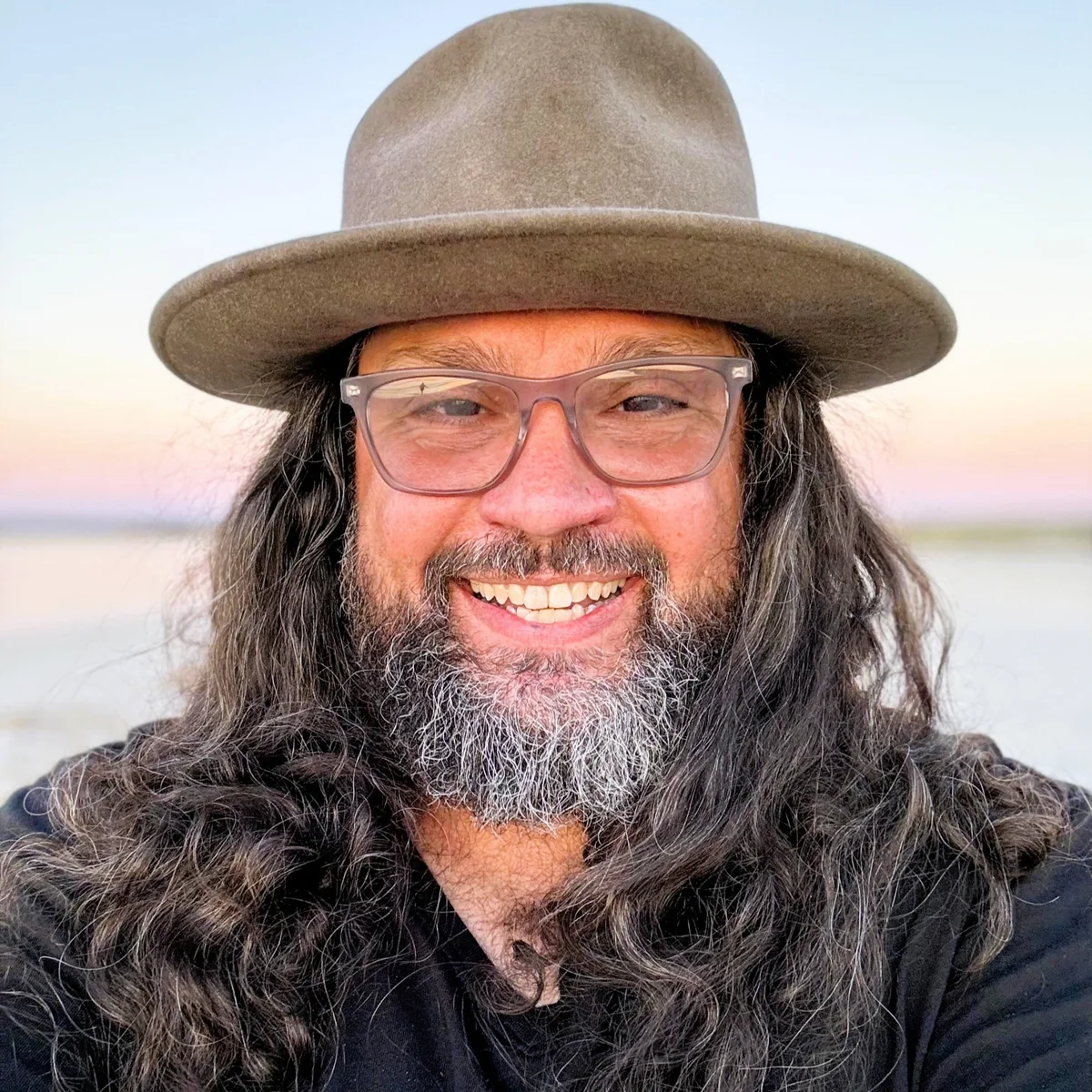 George Coldham smiling outdoors, wearing glasses and a broad-brimmed hat